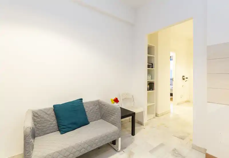 Soggiorno luminoso e minimal con un piccolo divano trapuntato, cuscino turchese e tavolino laterale; pareti bianche e pavimento in marmo creano un'atmosfera ariosa.