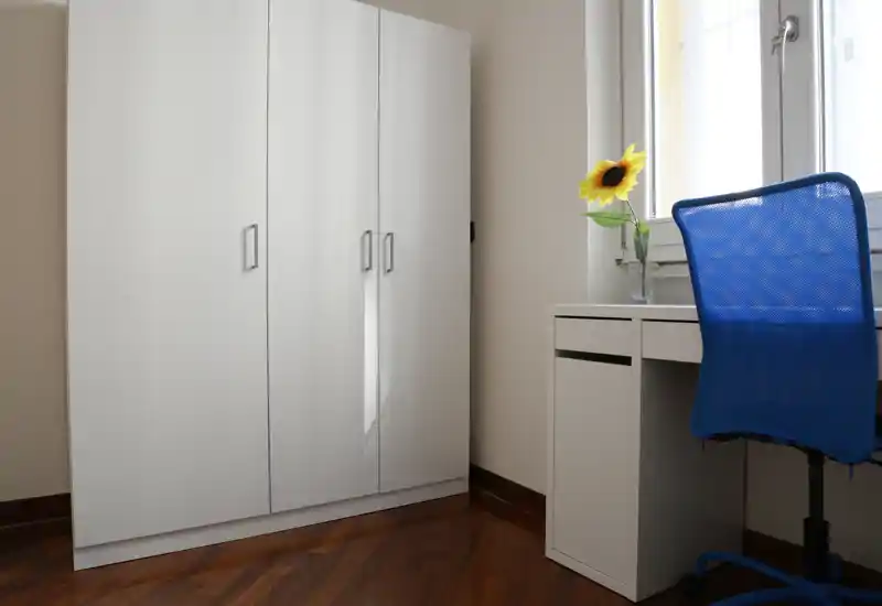 Small bedroom corner with a white wardrobe, a compact white desk by the window and a blue mesh chair; a single sunflower in a vase adds a cheerful touch and natural light brightens the space.