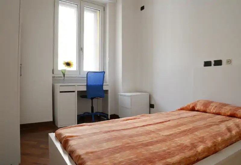 Bright, simple bedroom with a single bed dressed in warm orange linens, a small desk by the window and a compact bedside drawer — ideal for a student or single occupant.