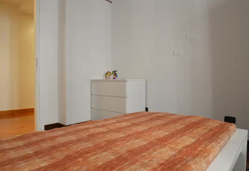 Bright, minimalist bedroom view featuring a large bed with warm orange bedding and a white chest of drawers against a neutral wall — simple, welcoming staging for a rental listing.