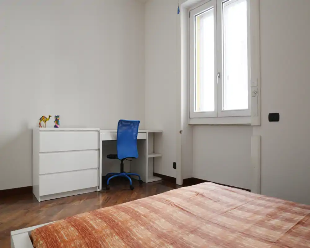 Camera da letto luminosa e minimal con letto parzialmente in primo piano, un comò bianco e una scrivania con sedia blu accanto a una grande finestra.