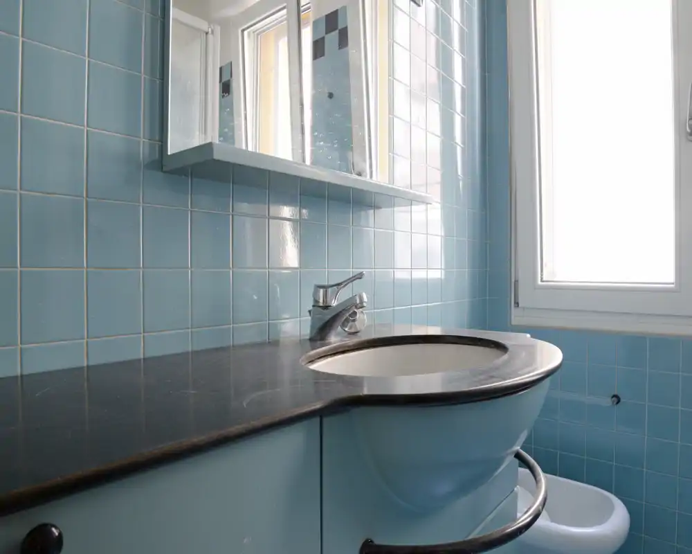Area lavabo compatta con pareti piastrellate turchesi, piano in nero lucido e lavabo circolare sotto un pensile a specchio; luce naturale intensa da una finestra vicina.