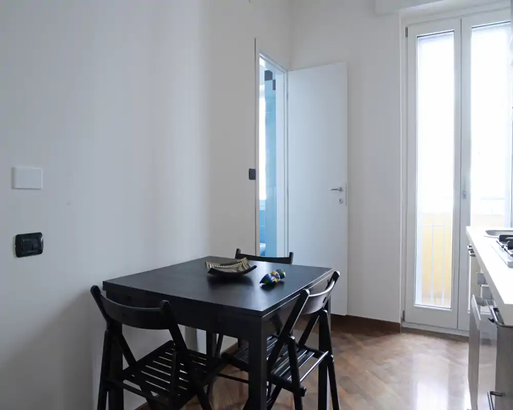 Angolo pranzo compatto con tavolo in legno scuro e quattro sedie pieghevoli, luce naturale dalla porta del balcone e un'estetica minimal pulita, ideale per un piccolo appartamento.