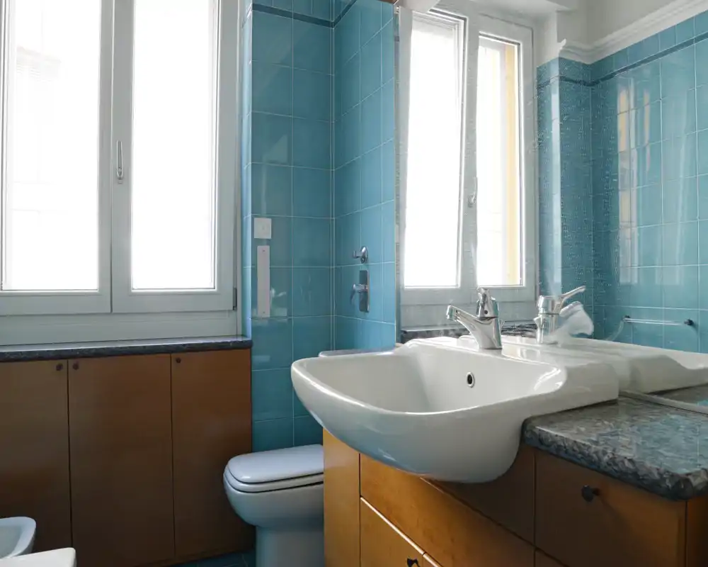 Bagno luminoso e ordinato con lavabo bianco, rubinetto moderno e piastrelle turchesi. La luce naturale dalla finestra mette in risalto il piano in granito e i mobili in legno.
