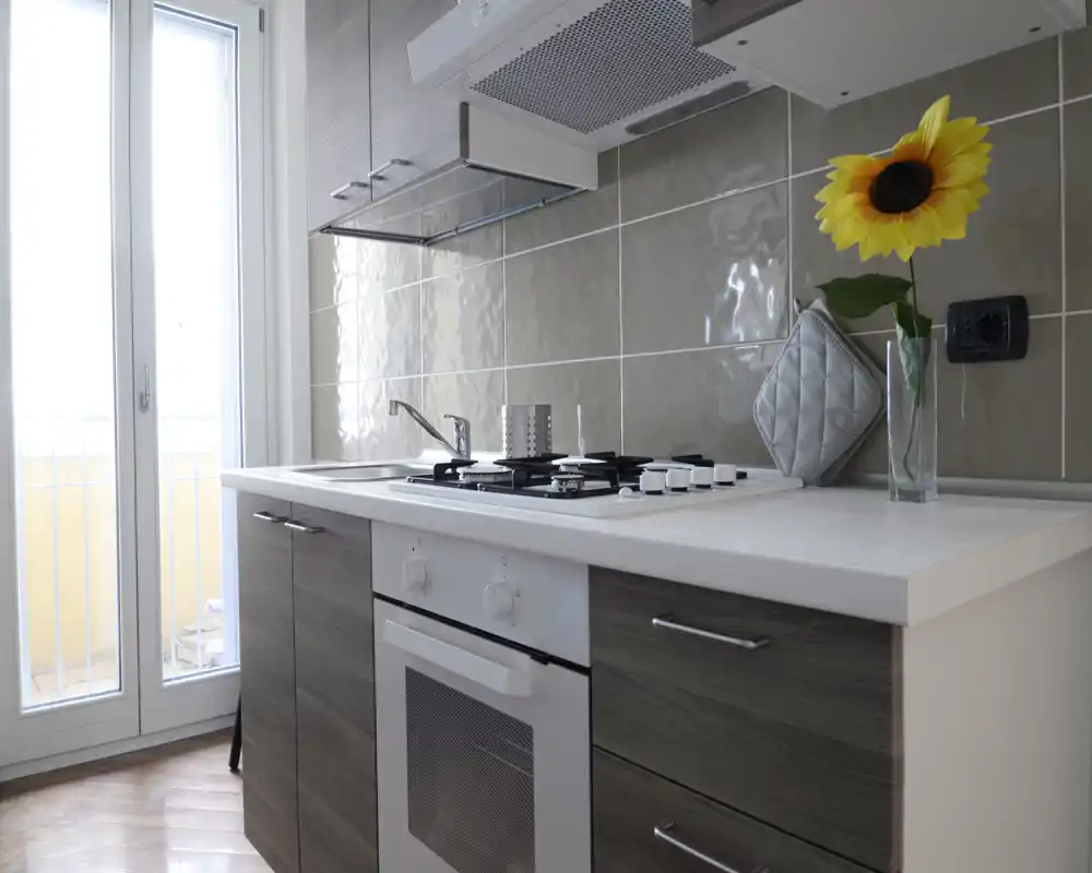 Cucina moderna e luminosa con forno incassato e piano cottura a gas, piano chiaro e porta finestra che garantisce luce naturale e accesso al balcone.
