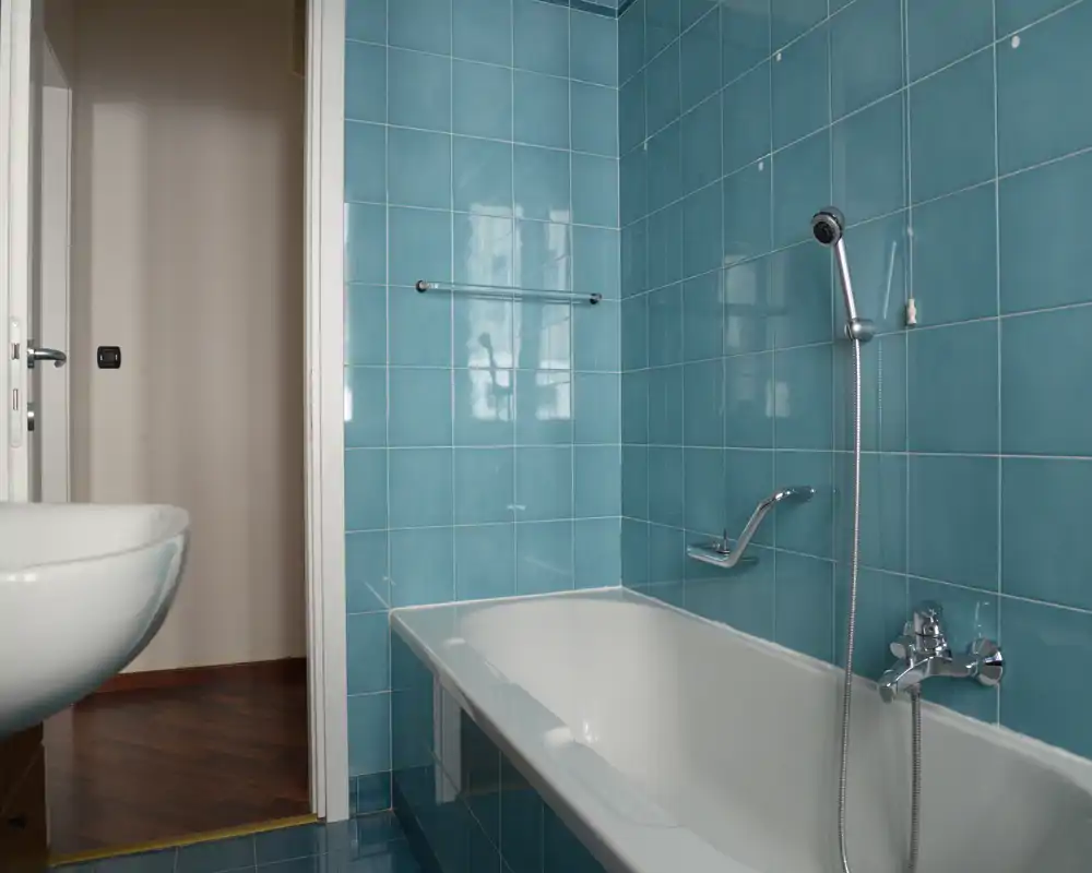 Bagno luminoso con pareti rivestite in piastrelle turchesi, vasca con doccia a mano e lavabo bianco parzialmente visibile; sanitari moderni e luce naturale che riflette sulle piastrelle.