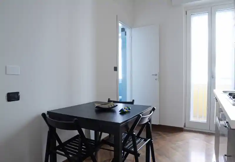 Angolo pranzo compatto con tavolo in legno scuro e quattro sedie pieghevoli, luce naturale dalla porta del balcone e un'estetica minimal pulita, ideale per un piccolo appartamento.