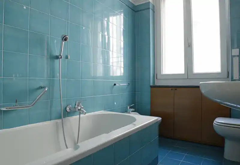 Bagno luminoso con vasca, doccia a mano, lavabo e WC; piastrelle turchesi e luce naturale da una finestra ampia danno una sensazione di pulizia e freschezza.