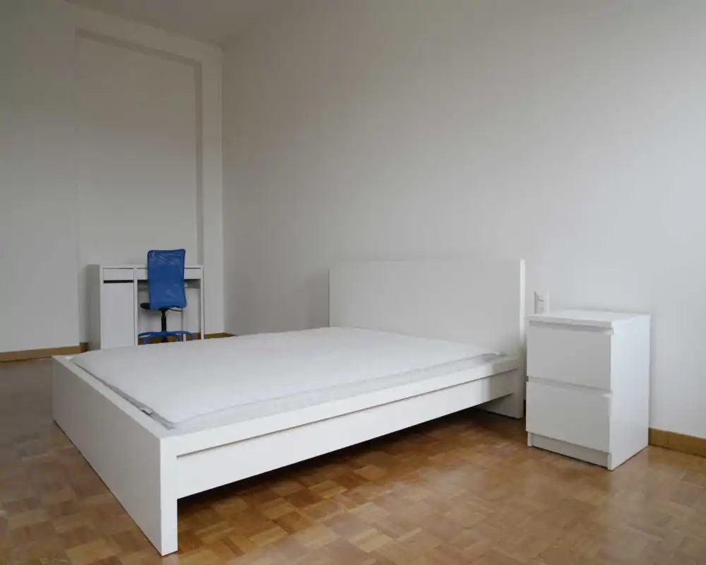 Chambre minimaliste avec un lit bas blanc, une table de chevet assortie et un petit bureau avec une chaise bleue ; parquet et murs neutres pour une ambiance moderne et apaisante.