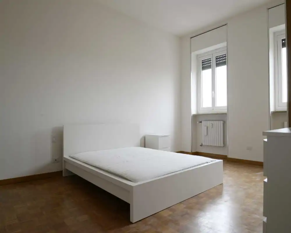 Chambre minimaliste et lumineuse avec un lit bas blanc sur parquet. Deux fenêtres apportent une lumière naturelle douce ; la pièce est épurée et prête à être aménagée.