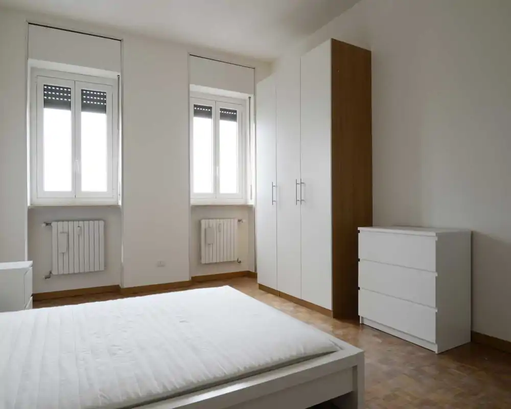 Chambre minimaliste avec un grand lit blanc au premier plan, deux fenêtres lumineuses et une armoire et commode blanches assorties — propre, aérée et épurée.