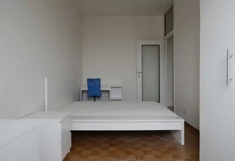 Chambre minimaliste et lumineuse avec cadre de lit blanc, matelas et un petit bureau avec chaise bleu ; parquet propre et murs neutres offrant un espace calme et simple.