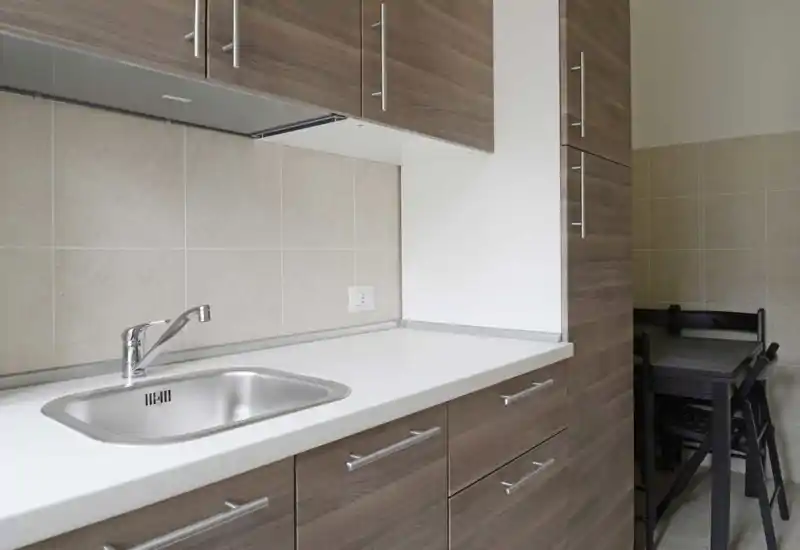 Cucina moderna compatta con lavello in acciaio, mobili effetto legno e piano bianco pulito — luminosa e ordinata.