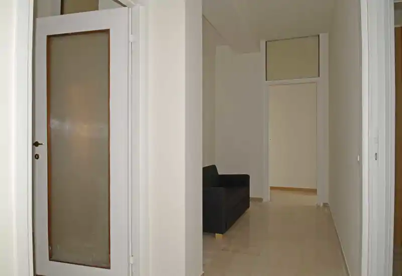 Espace de vie étroit ou couloir avec un petit canapé sombre contre un mur clair ; sol carrelé et porte translucide offrant un espace simple et fonctionnel.