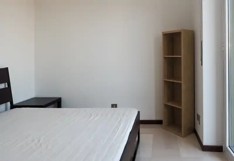 Chambre minimaliste avec cadre de lit double et matelas, table de chevet et étagère haute ouverte ; lumière naturelle abondante et murs neutres pour une ambiance reposante.
