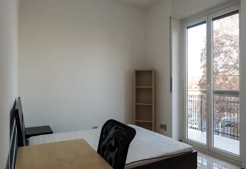 Chambre minimaliste avec lit simple, chaise de bureau et étagère. Grande porte-fenêtre donnant sur le balcon, apportant lumière naturelle et une jolie vue.
