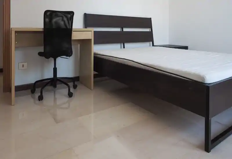 Chambre simple et moderne avec un cadre de lit en bois foncé et un matelas propre. La pièce comprend un petit bureau et une chaise de bureau, sol carrelé poli et murs neutres — lumineuse et minimaliste.