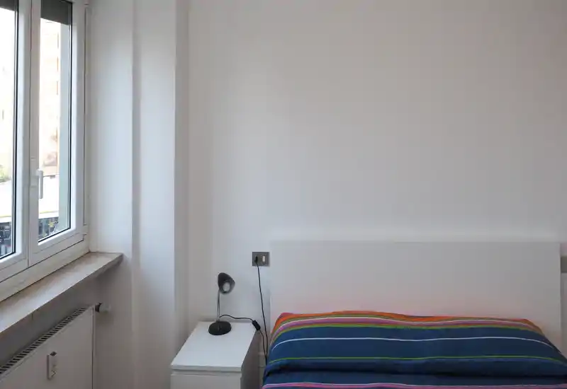 Chambre minimaliste et lumineuse avec tête de lit blanche, couette à rayures et table de chevet avec lampe près d'une grande fenêtre laissant entrer la lumière naturelle.