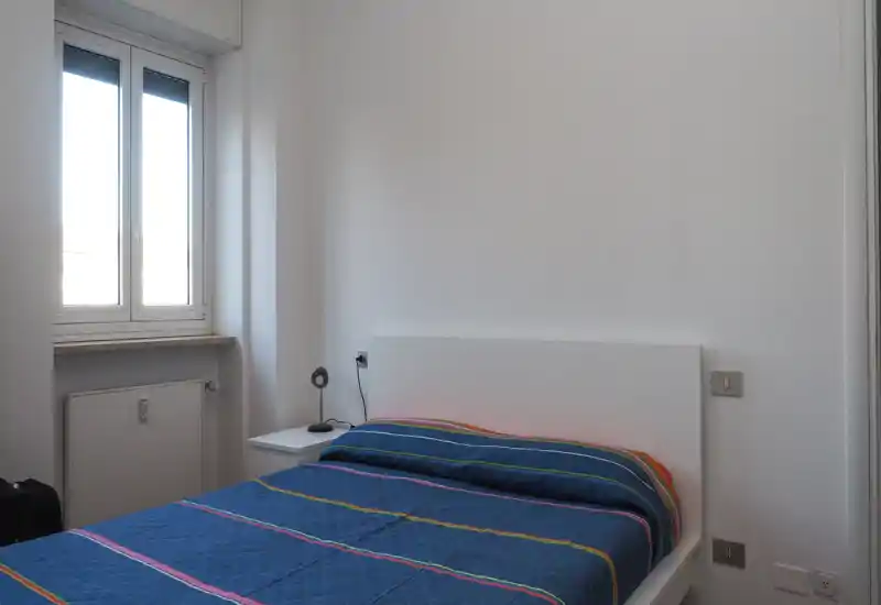 Chambre lumineuse et minimaliste avec un lit double, couette bleu à rayures et une table de chevet avec une petite lampe. La lumière naturelle de la fenêtre rend la pièce aérienne et soignée — idéale pour un séjour confortable.