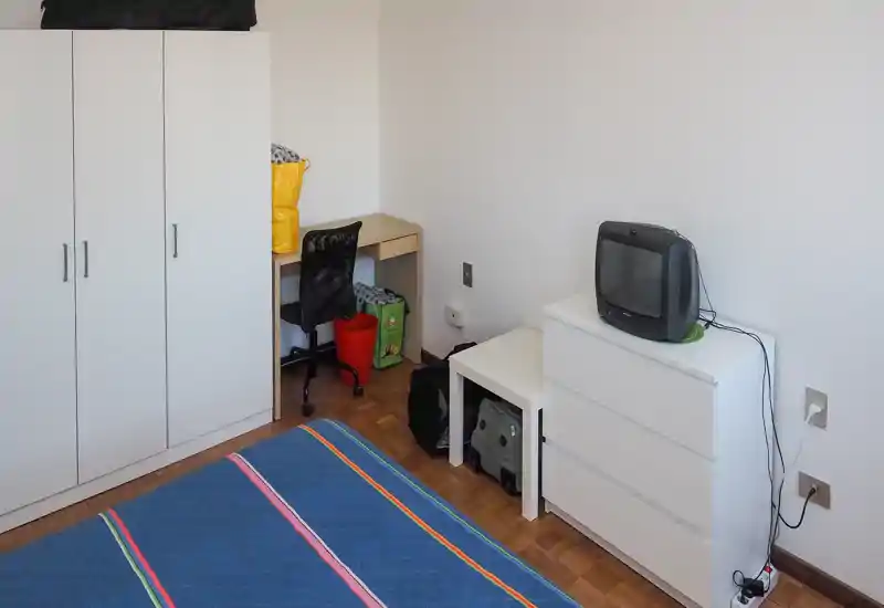 Chambre compacte et bien rangée avec un lit double au premier plan, une armoire blanche, une commode et un petit bureau avec chaise. Mobilier simple et fonctionnel, parquet et murs neutres, adaptée aux locations de courte durée.