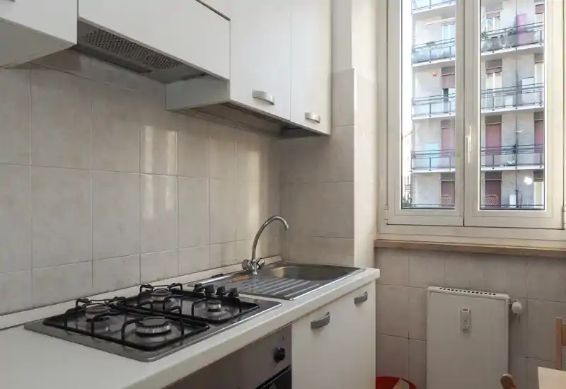 Cuisine compacte et fonctionnelle avec plaque à quatre feux, évier en inox et grande fenêtre apportant la lumière naturelle. Carrelage neutre et meubles blancs pour un espace de cuisson pratique et épuré.