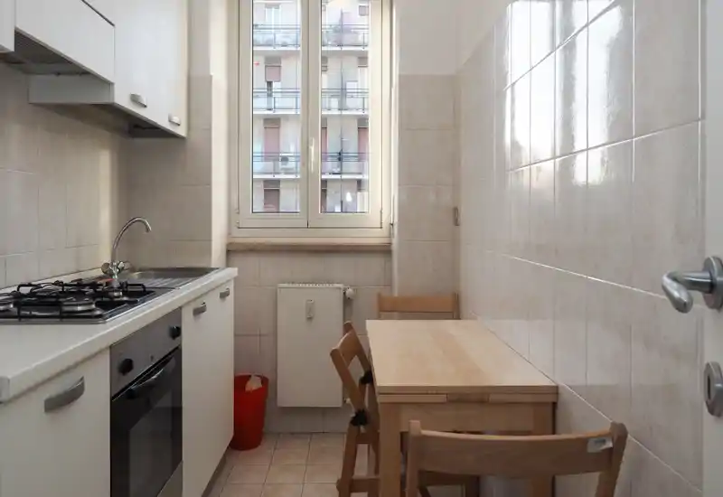 Cuisine en enfilade compacte avec cuisinière à gaz, évier et petite table en bois avec chaises près d'une fenêtre lumineuse. Carrelage propre et mobilier sobre confèrent un espace fonctionnel et bien éclairé, idéal pour la location courte durée.