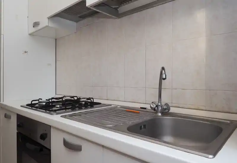 Cuisine compacte et fonctionnelle avec évier en acier inoxydable, égouttoir intégré et plaque à gaz. Meubles blancs et crédence carrelée pour un espace de cuisine pratique, adapté aux studios ou locations courtes durées.