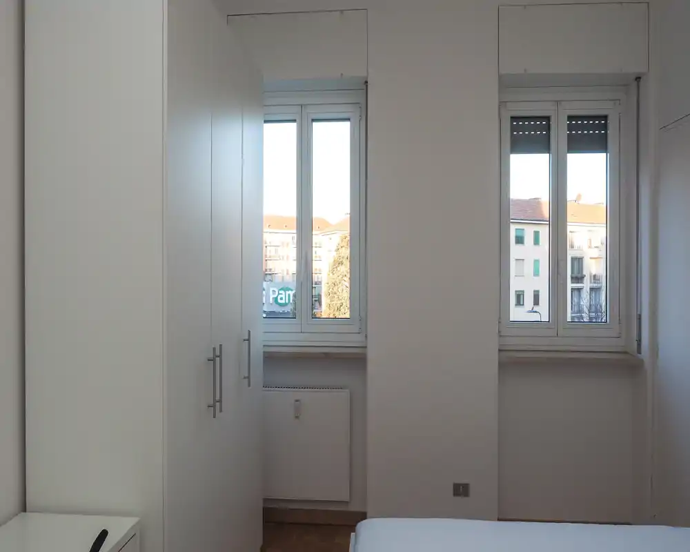 Minimal, bright bedroom corner with two large windows, a white wardrobe and a partially visible bed; clean, modern and well lit by daylight.