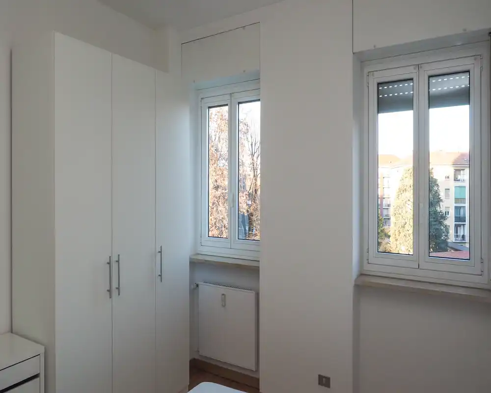 Small, bright bedroom corner with white wardrobe and two windows letting in natural light; the bed is only partly visible, giving a clean, minimal look.