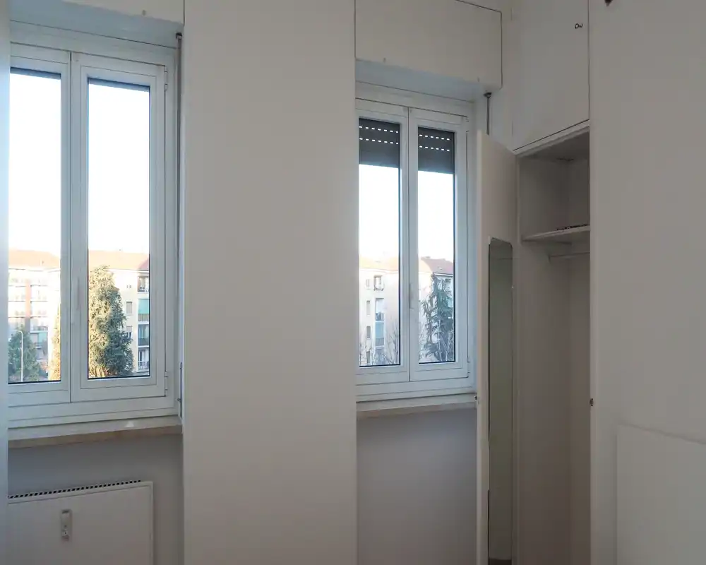 Small neutral interior space with two large windows and fitted storage. Bright natural light and a simple radiator beneath the windows; suitable for a hallway or small entrance area.