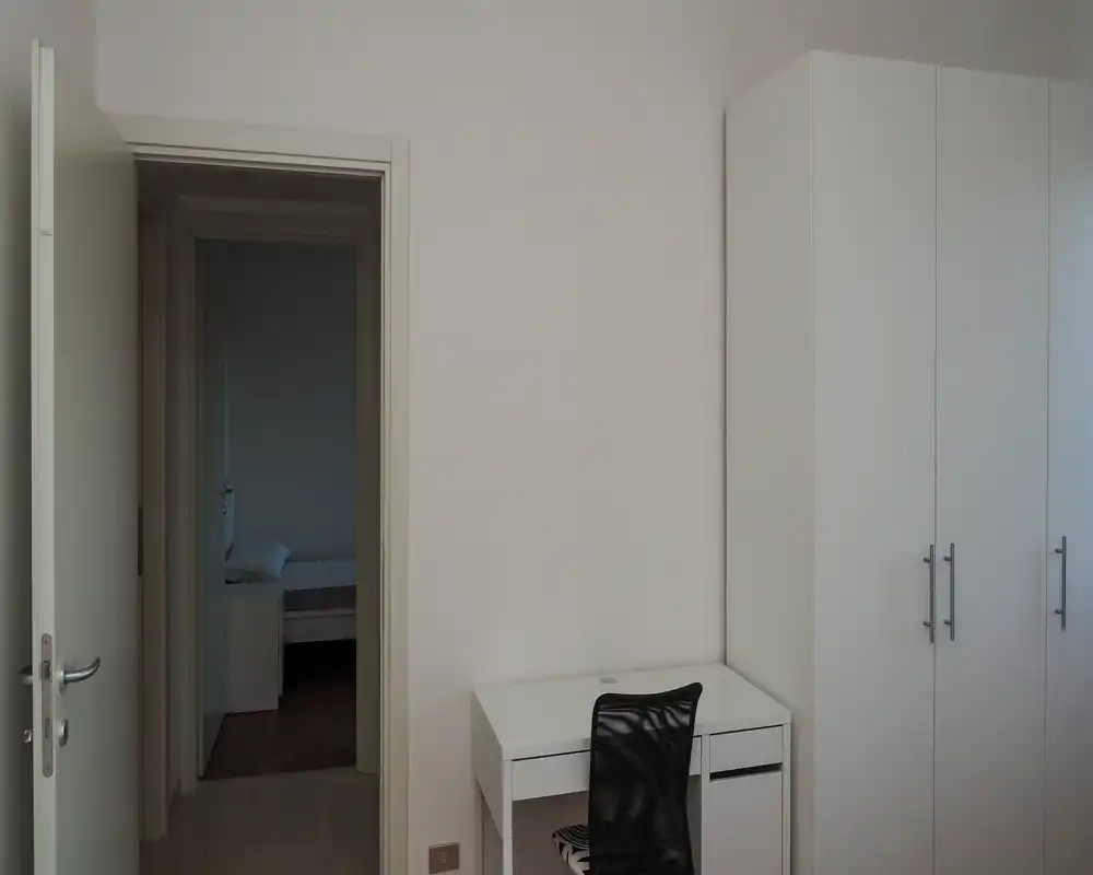 Quiet minimalist bedroom view: an open door reveals a simple bed and nightstand in an adjacent room; foreground shows a white desk, chair and wardrobe.