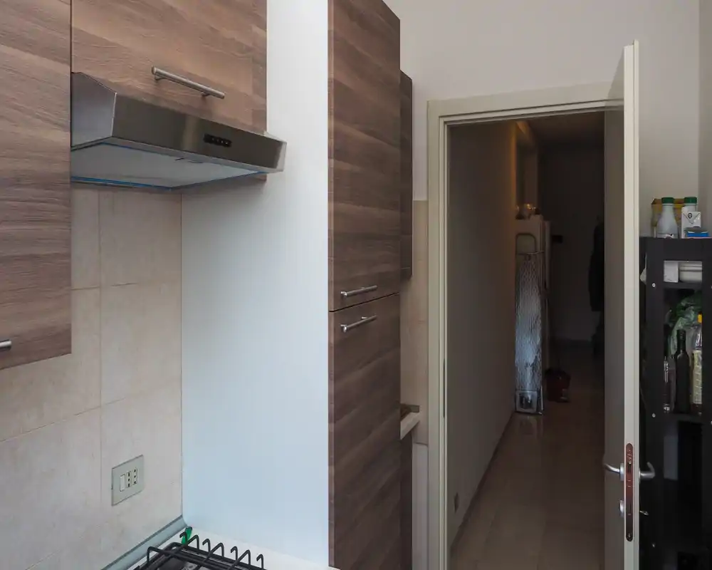 Compact modern kitchen corner with wood-effect cabinets, stainless steel extractor hood and a gas hob visible in the foreground. The layout appears functional with neutral tiles and organized storage.