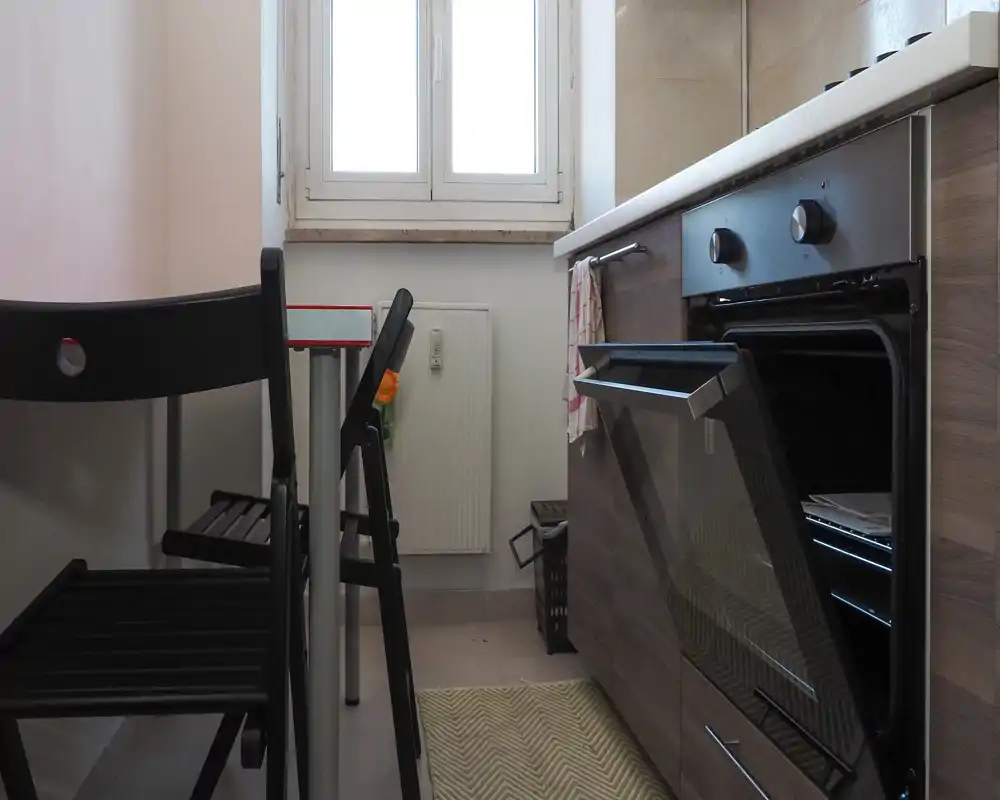 Compact kitchen with a built-in oven, countertop and small dining chairs next to a window; tidy and functional space ideal for a small apartment.