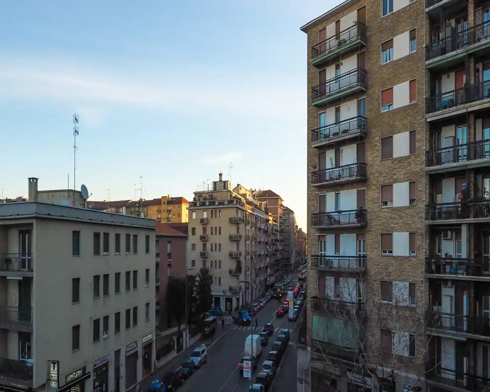 Street view from a balcony or window showing an urban residential street with mid-rise apartment buildings and parked cars, captured in soft evening light.