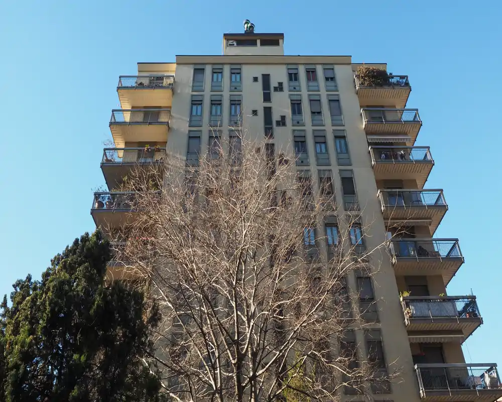 Exterior view of an apartment building with multiple balconies taken from street level; clear blue sky and leafless tree in the foreground.