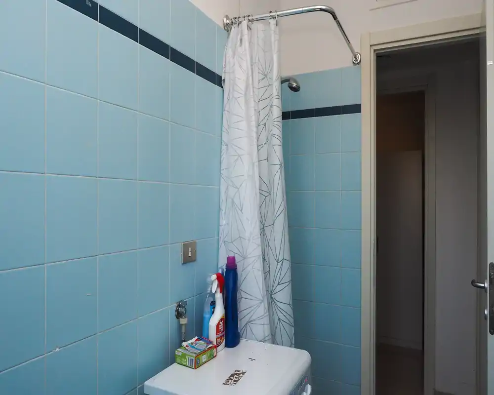 Small bathroom with light-blue tiled walls, a shower area with curtain and a top-mounted showerhead, and cleaning products placed on a small surface.