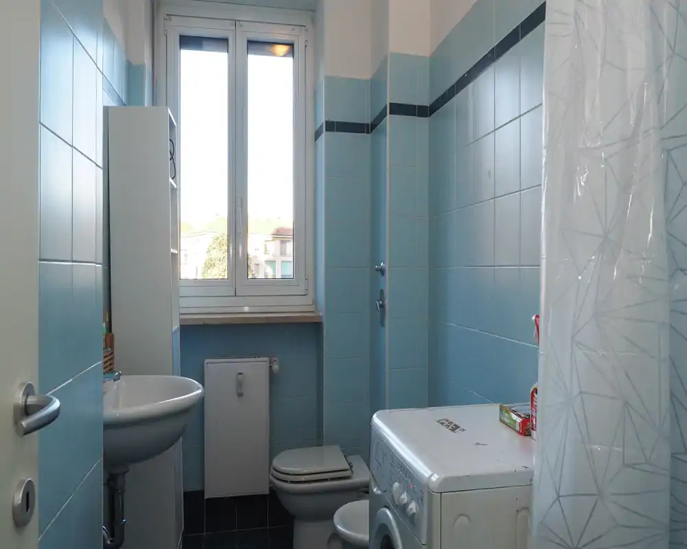 Compact bathroom with blue wall tiles, a sink, toilet, shower area with curtain and a washing machine under pleasant natural light from the window.