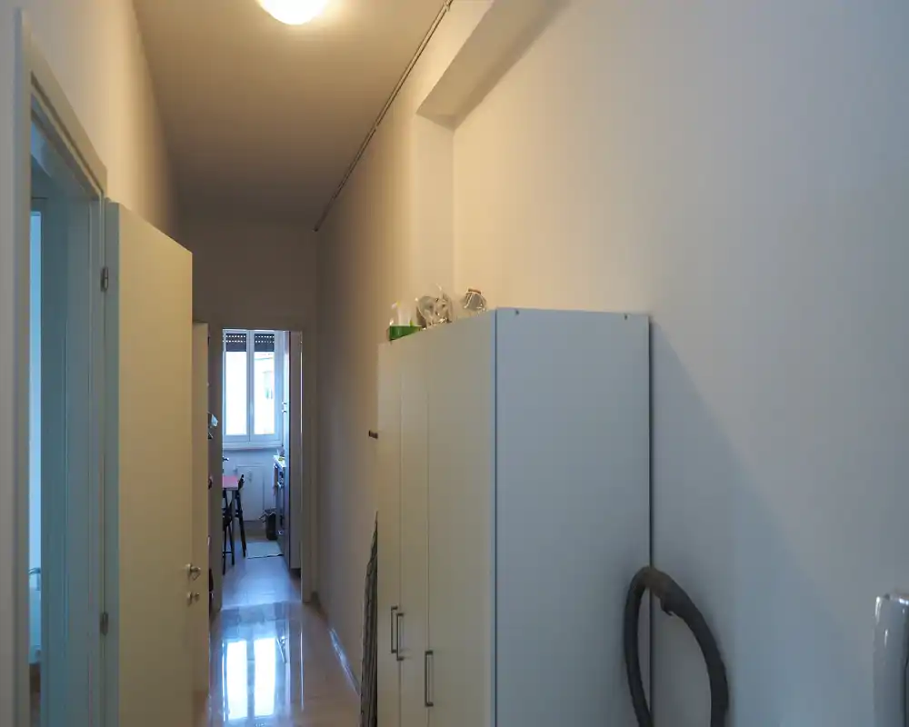 Narrow entrance/hallway with built-in storage cabinets, neutral walls and tiled floor, leading toward a lit room with a window.