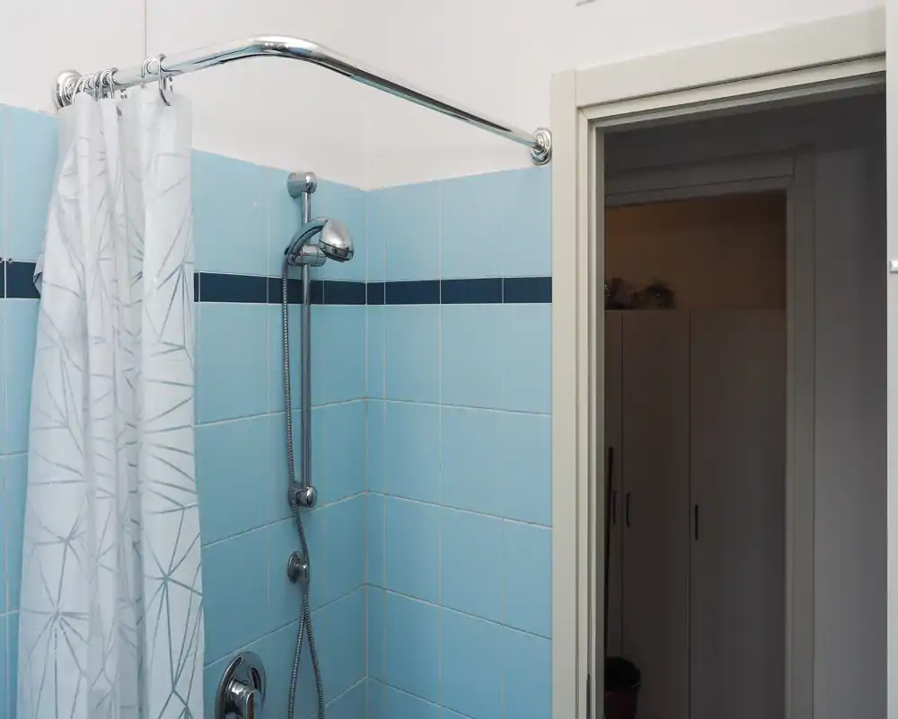 Clean, tiled bathroom corner with a wall-mounted adjustable shower head and a patterned shower curtain; pale blue tiles and chrome fixtures give a fresh, functional look.