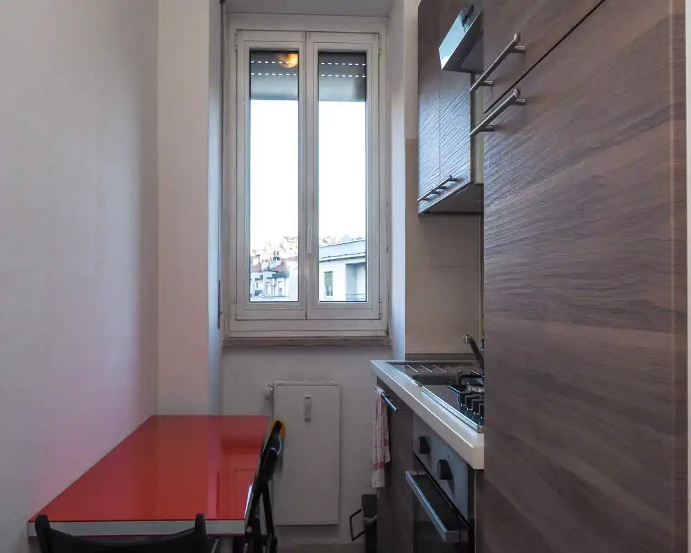 Compact fitted kitchen with a gas hob, oven and a small red dining table by a bright window — ideal for a city apartment.