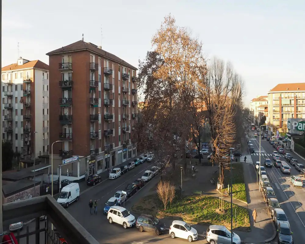 City street view from a balcony overlooking a small park and surrounding apartment buildings, with parked cars and tree-lined sidewalks in warm late-afternoon light.