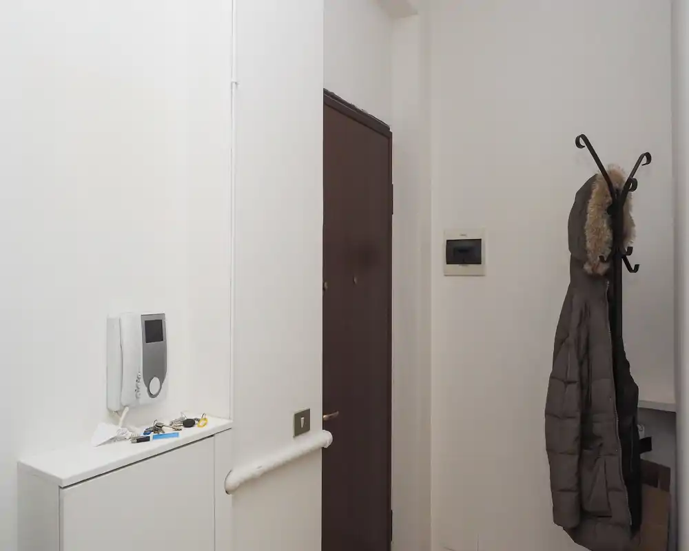 Compact, well-lit entryway with a wall-mounted intercom, small shelf with keys and a coat rack holding a winter jacket — ideal as a practical apartment entrance.