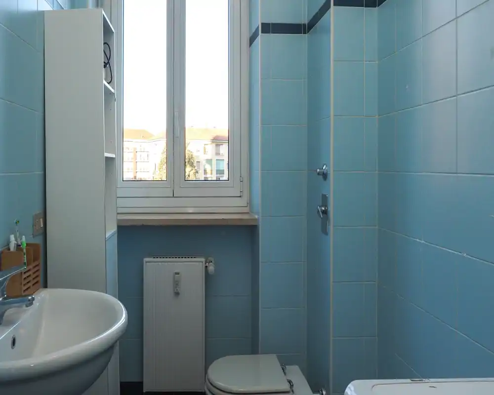 Small, tidy bathroom with light blue tiles, a window providing natural light, a sink, toilet and radiator, ideal for a compact apartment.