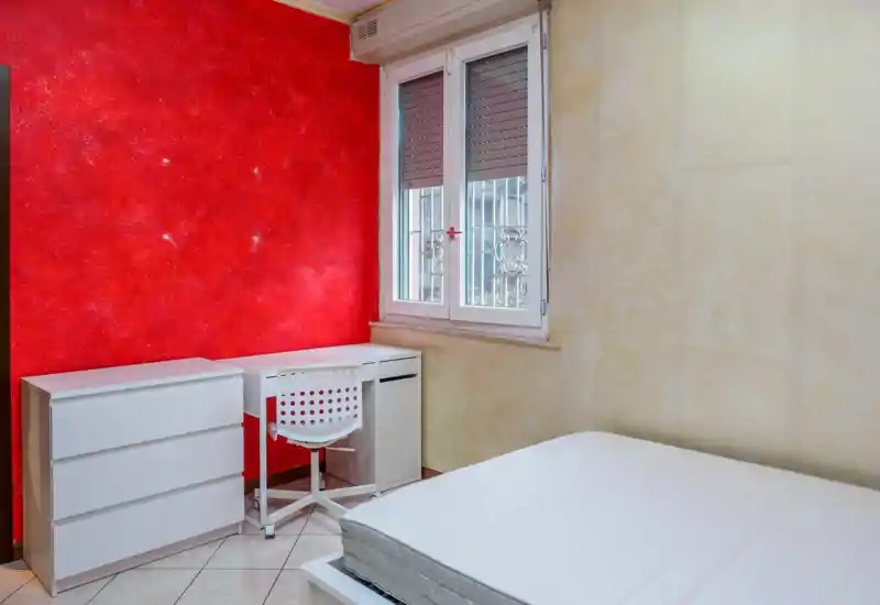 Camera da letto luminosa con materasso bianco in primo piano, una scrivania compatta e un comò bianchi contro una parete rossa testurizzata e una finestra che fornisce luce naturale.