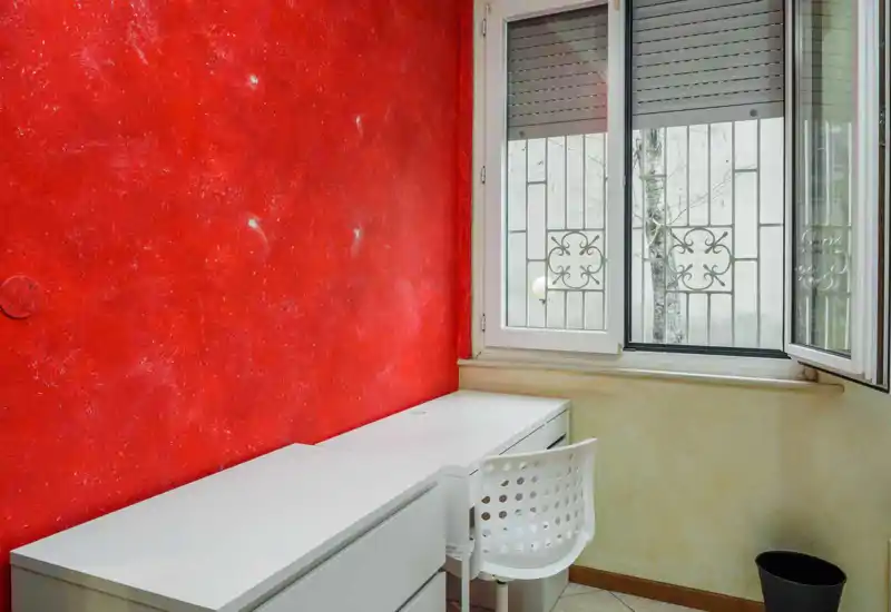 Angolo studio compatto con scrivania e sedia bianche vicino alla finestra; la parete rossa texturizzata dà personalità e la finestra offre luce naturale.