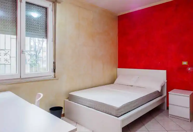 Camera da letto luminosa e semplicemente arredata con letto matrimoniale su struttura bianca, due cuscini, comodino bianco e una scrivania vicino alla finestra; parete rossa a contrasto.