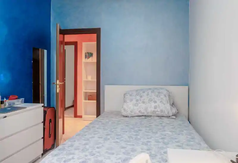 Camera da letto luminosa e semplice con letto a motivi davanti a una parete azzurro chiaro, un comò bianco e una porta aperta verso il corridoio. La stanza è ordinata e funzionale con luce naturale soffusa.