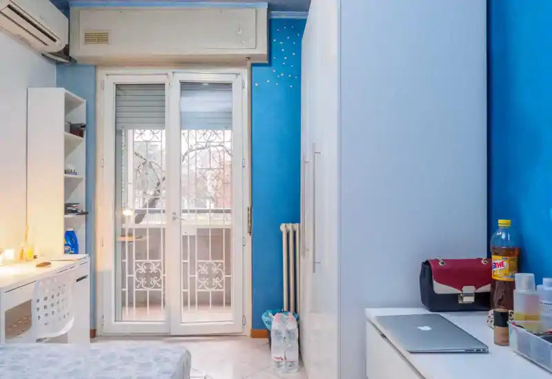 Camera da letto luminosa e ordinata con letto matrimoniale in primo piano, scrivania e scaffali a sinistra e porta finestra che illumina naturalmente.