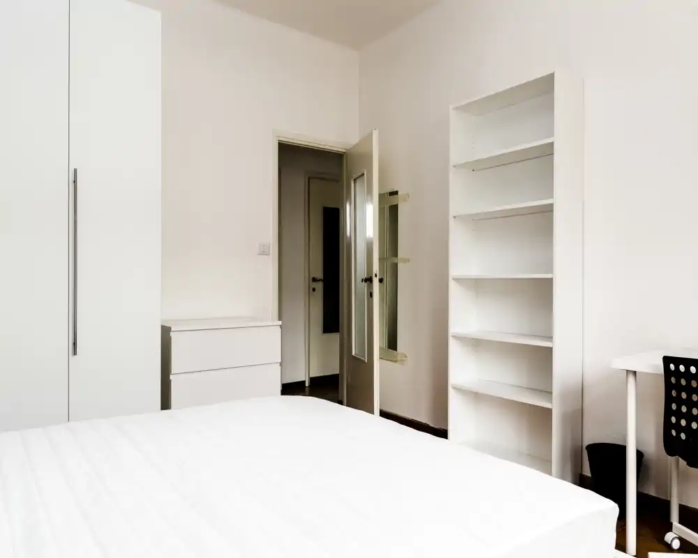 Dormitorio minimalista con colchón blanco grande, armario, estantería y un pequeño escritorio; tonos neutros y luminosos que transmiten tranquilidad.
