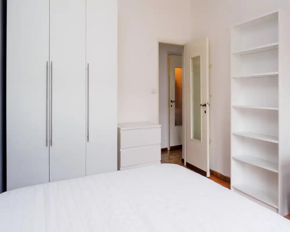 Dormitorio luminoso y minimalista con una gran cama blanca en primer plano, armario empotrado, cómoda y estanterías — ambientación moderna y limpia, perfecta para alquileres.